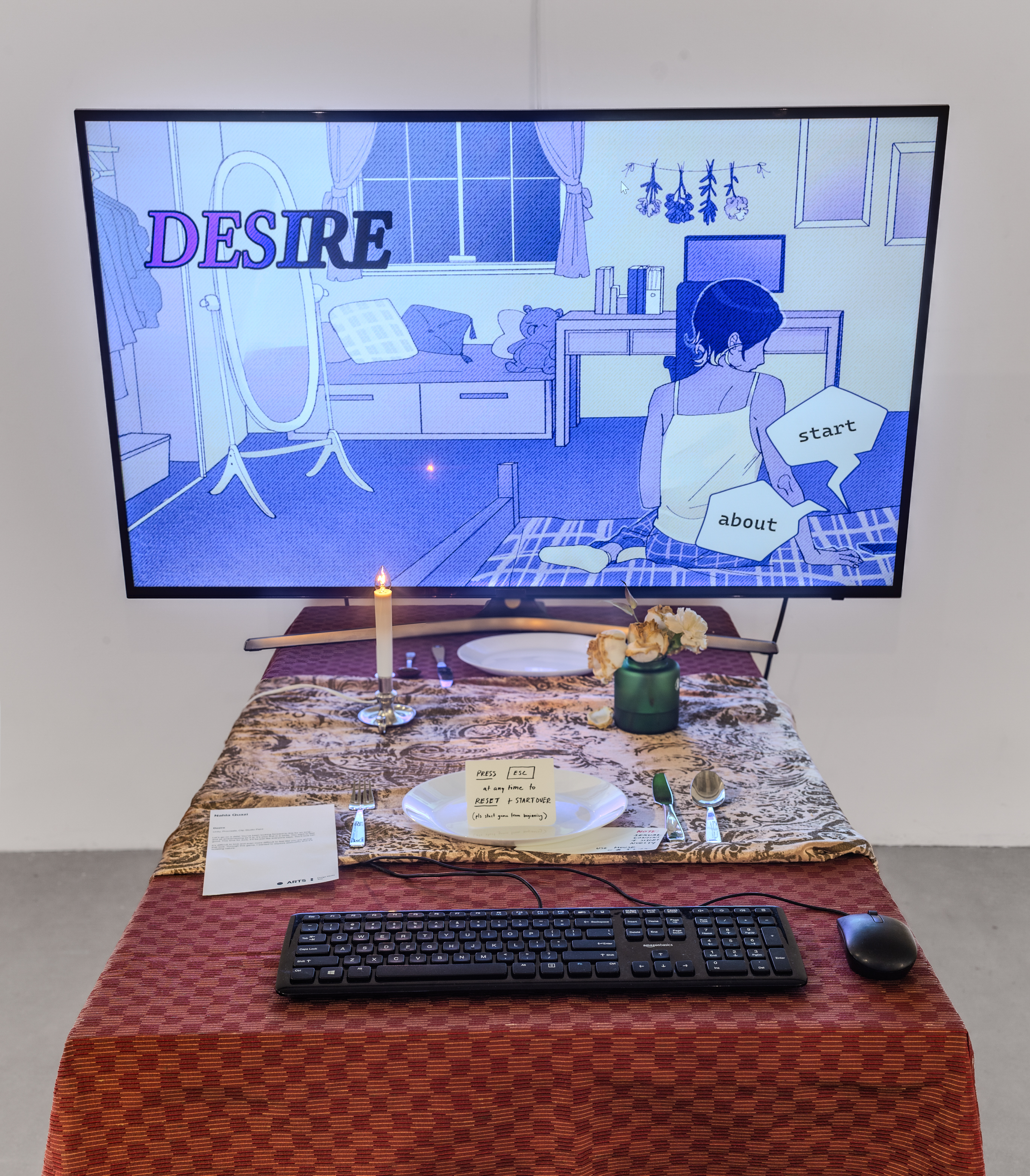 An upholstered chair sits in front of a table with tablecloth. On top of the table is a keyboard, a computer mouse, another table runner, a glass case with two flowers, a fake wax candle, and a television. On the TV is the start screen of an interactive game.